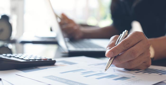 Close de uma mão escrevendo sobre gráfico com uma calculadora ao fundo